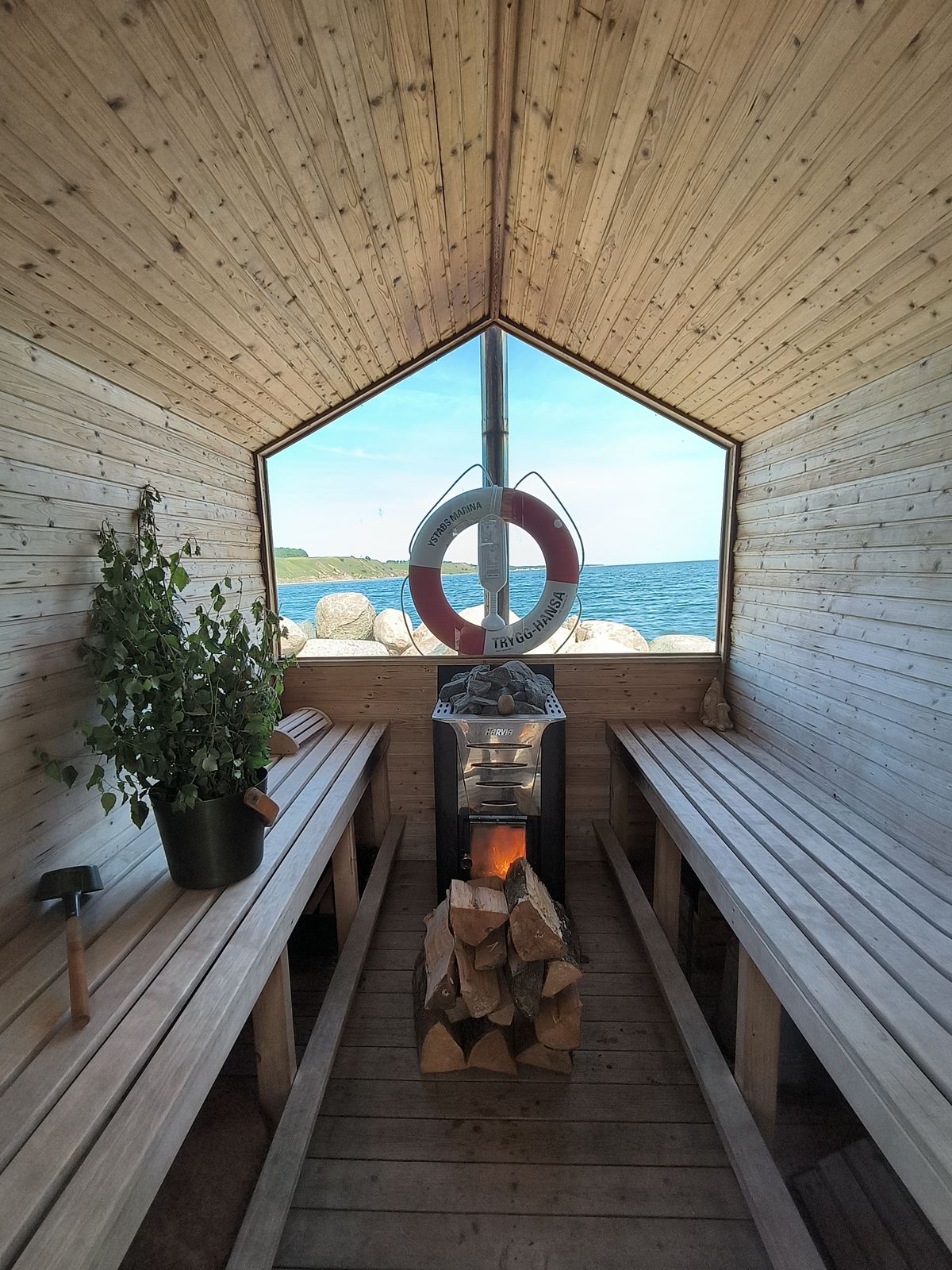 Ljust bastuinteriör med utsikt över havet, vedeldad kamin och björkvihta.
